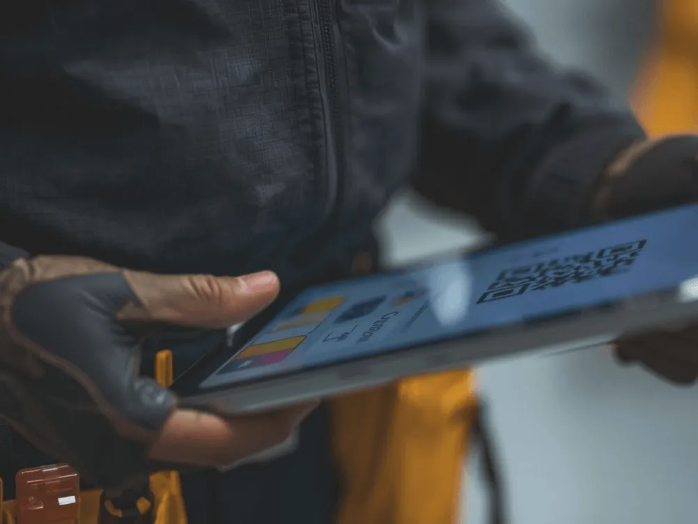 A field service professional wearing protective gloves and a tool belt using a digital tablet to display a QR code for on-site business operations.