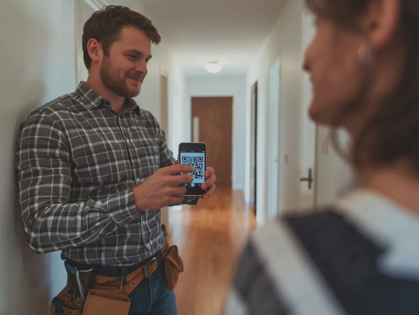 A smiling service technician in a plaid shirt and tool belt showing a smartphone with a QR code to a customer in a home hallway.